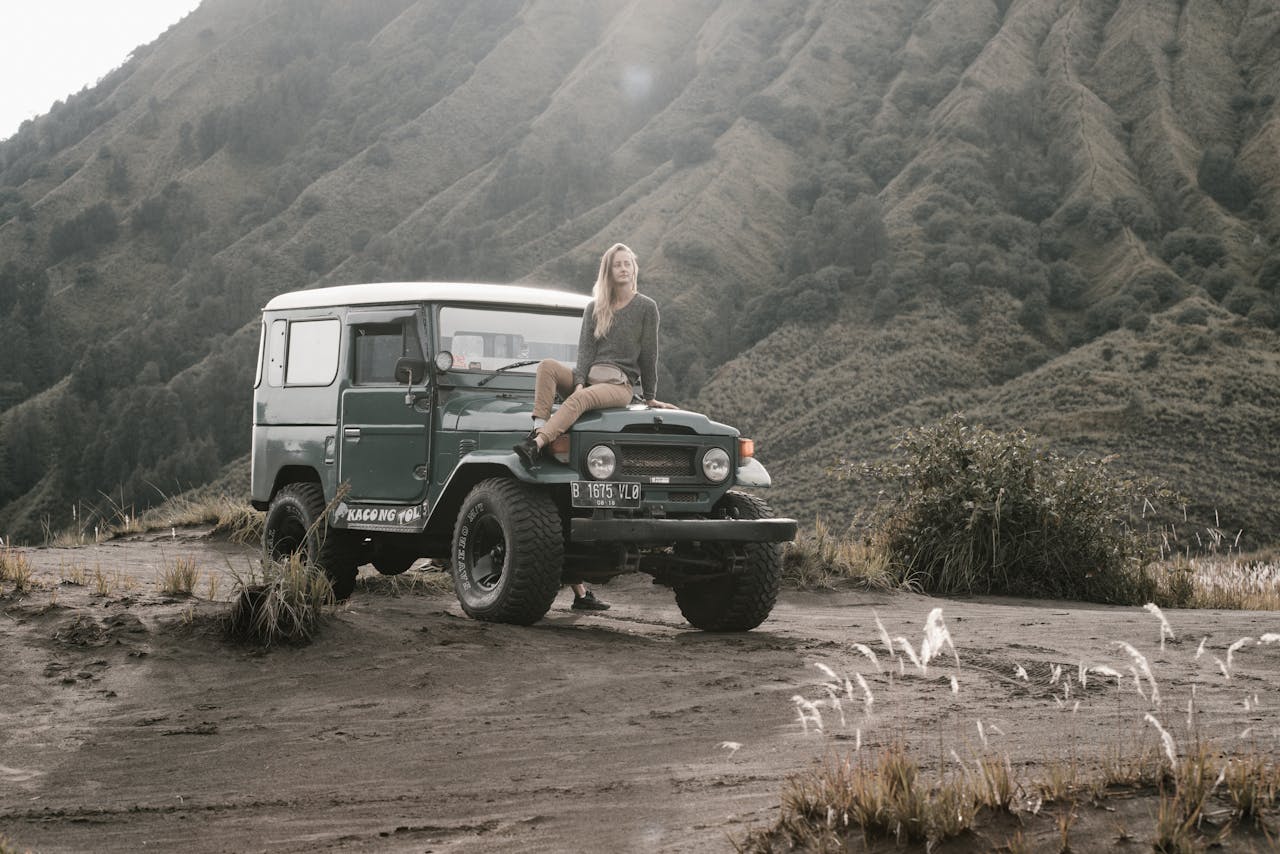 A woman sits on a jeep in a rugged mountain landscape.